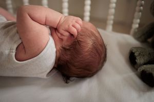 Newborn baby lying in a white crib with hand raised to head, with a plush toy nearby.