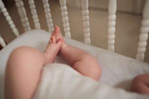 Newborn baby's tiny bare feet and legs resting against white crib mattress between crib slats.