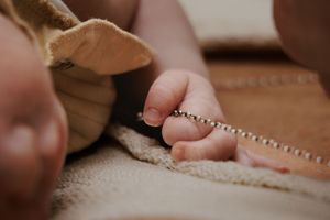Newborn baby hand grasping a delicate beaded bracelet on a soft brown blanket.