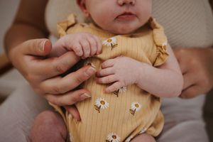 Newborn baby in yellow daisy romper grasping parent's finger with tiny hand.