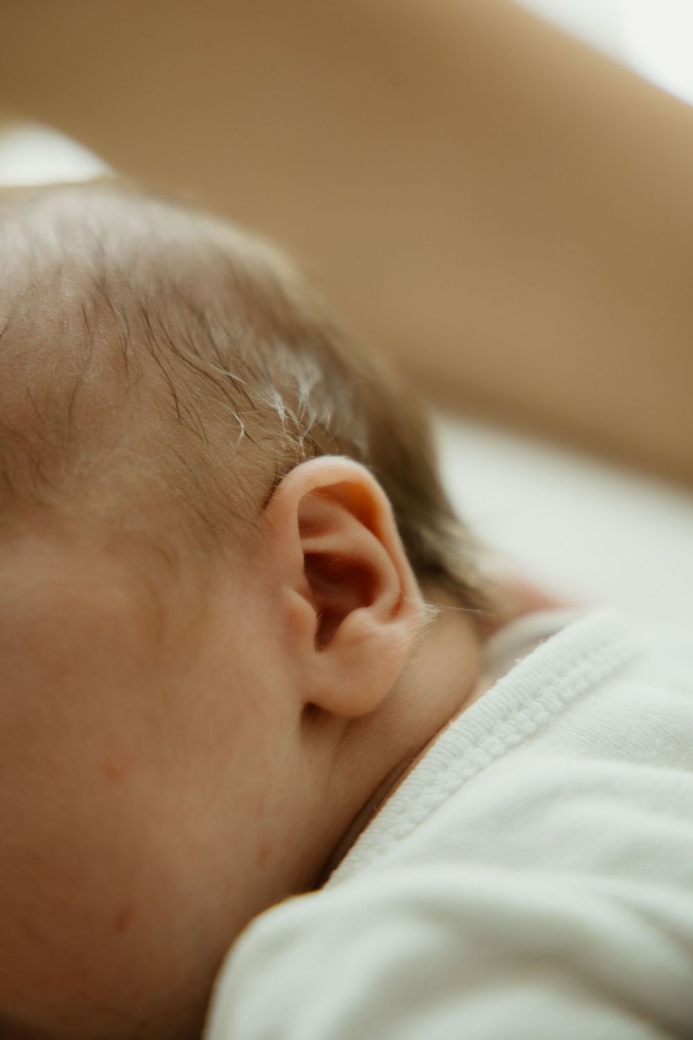 Close-up of a sleeping newborn baby's ear and dark hair, wearing a white onesie.