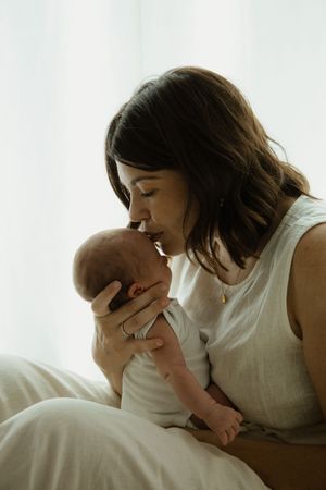 A mother tenderly kisses her newborn baby while sitting by a bright window in soft natural light.
