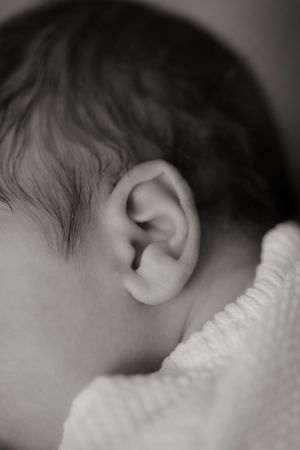 Black and white close-up photo of a newborn baby's ear and dark hair wrapped in a white blanket.