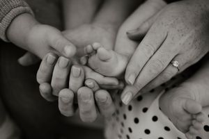 Black and white photo of family hands cradling a newborn baby's tiny feet, with a diamond engagement ring visible.