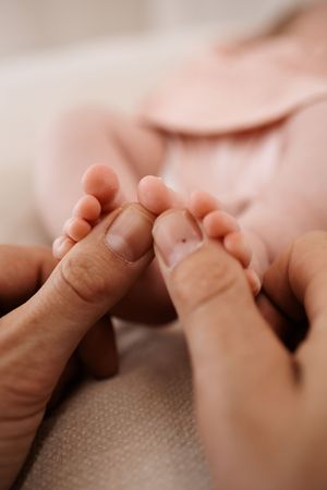 Adult hands gently holding tiny newborn baby feet in soft pink outfit.