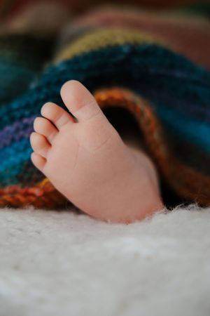 Tiny newborn baby foot peeking out from under a colorful striped blanket on a white fluffy surface.