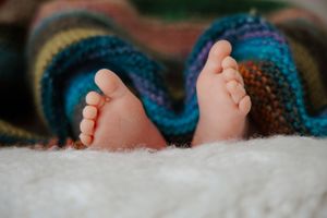 Close-up of newborn baby's two tiny feet emerging from a colorful knitted blanket on a soft white surface.