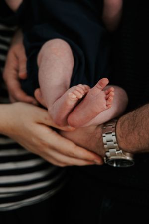Parents' hands gently supporting newborn baby's tiny feet, with father's silver watch visible in warm-toned photo.