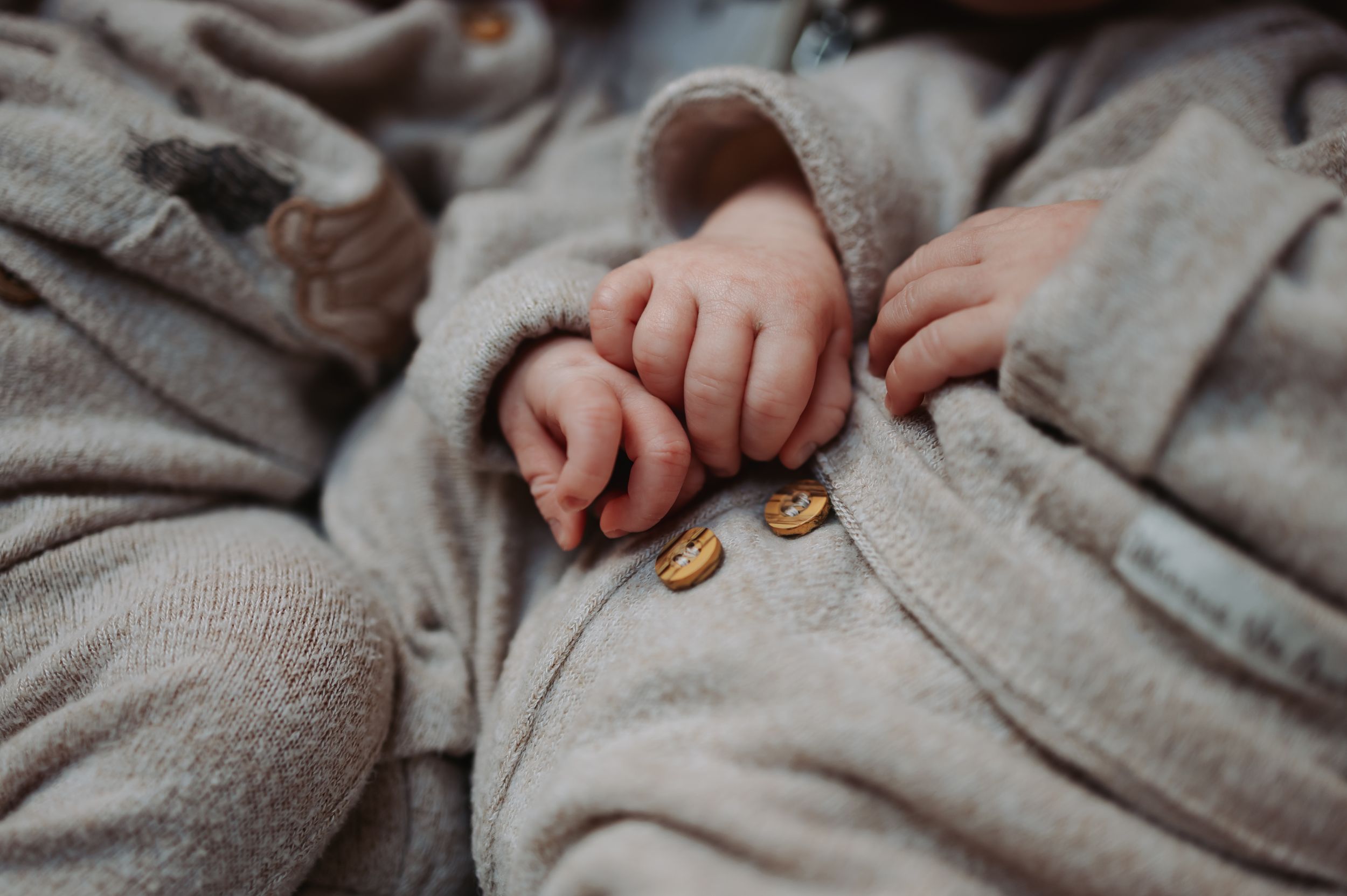 Close-up of a newborn baby's tiny hands resting on a soft beige fleece onesie with gold buttons.