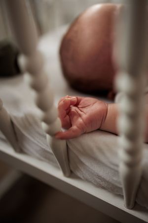 Newborn baby hand resting on white crib mattress between white wooden crib bars.