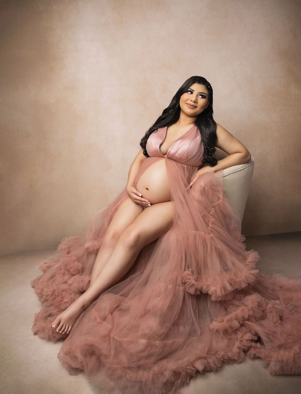 A glamorous maternity portrait in flowing pink tulle gown against neutral background.