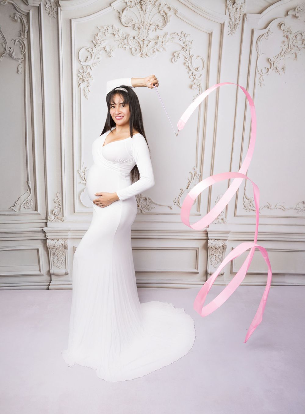 Maternity portrait with pink ribbon in elegant white dress against ornate wall backdrop.