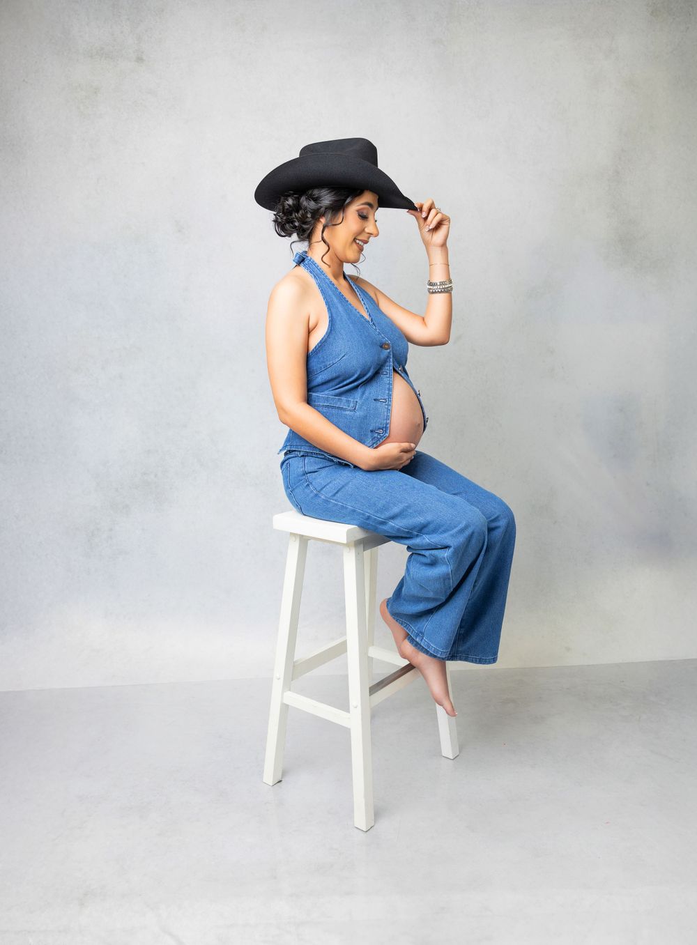 A side view maternity photo of woman in denim and black cowboy hat seated on white stool.