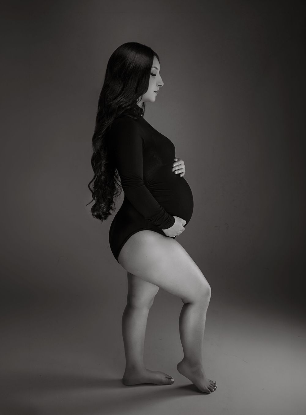 A black and white image of a pregnant woman with long dark hair posing in a bodysuit.