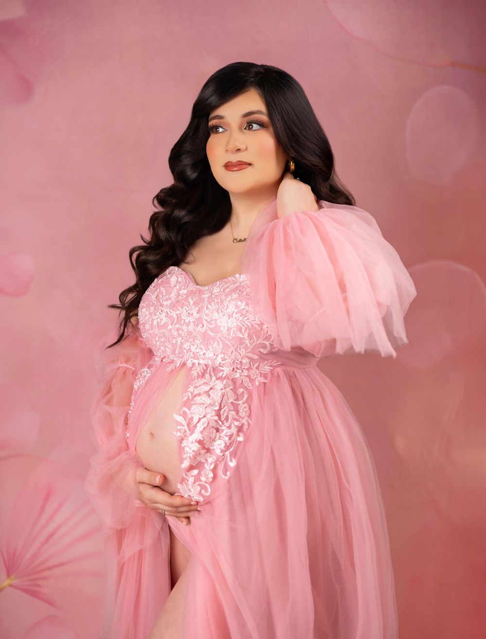 Pregnant woman in pink tulle maternity gown posing gracefully against a soft floral pink studio background.
