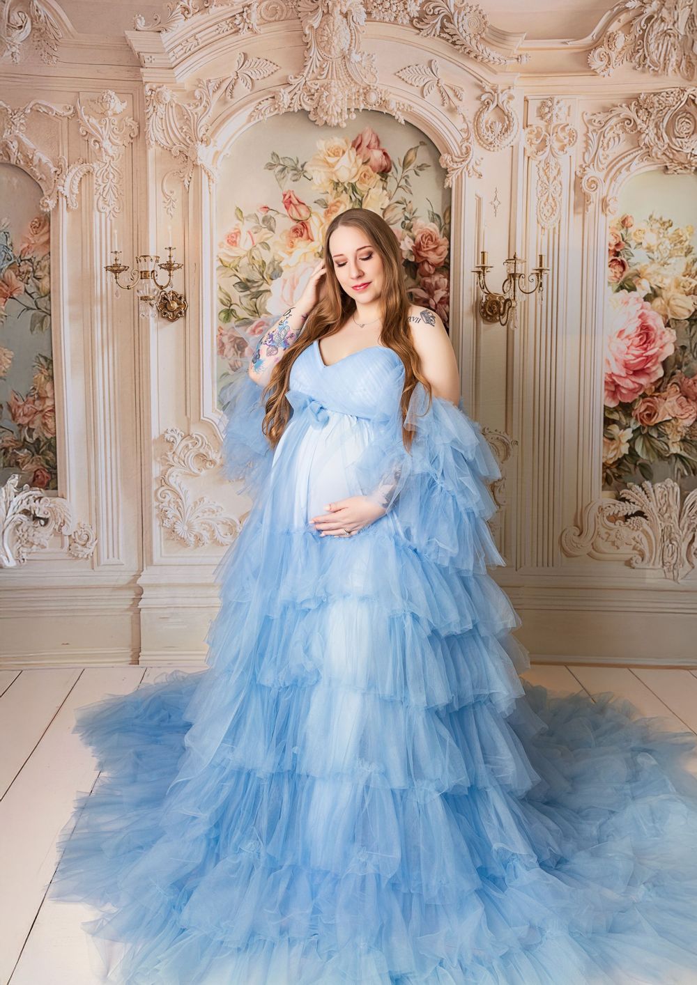 Ethereal light blue tulle maternity gown with tiered ruffles photographed against ornate rococo wall with floral details.