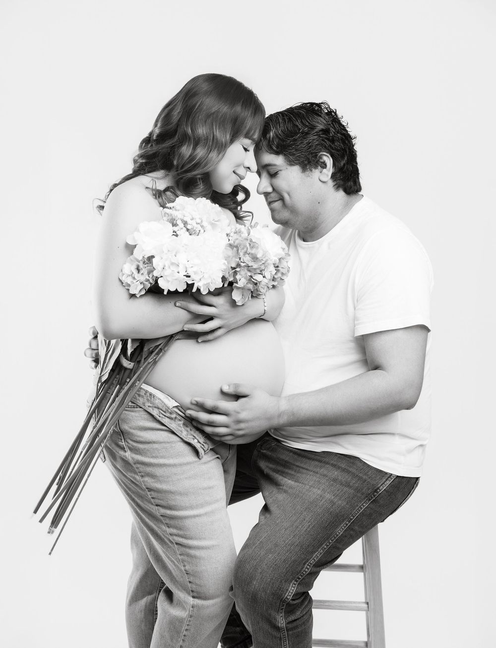 Black and white maternity photo of couple, woman holding flowers over baby bump, man tenderly touching her belly.