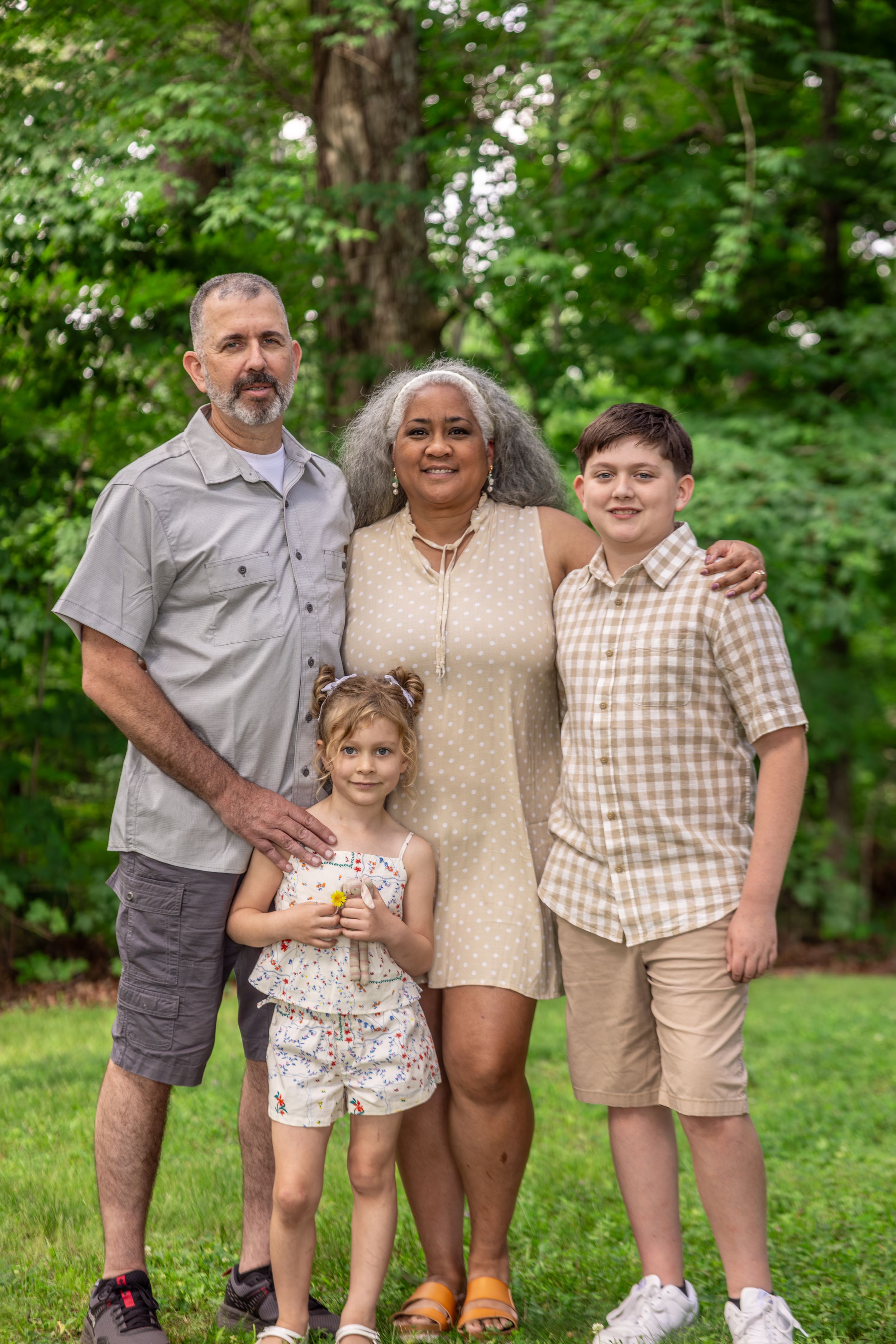 Multigenerational family of four posing outdoors in a lush green park, smiling together on a sunny day.