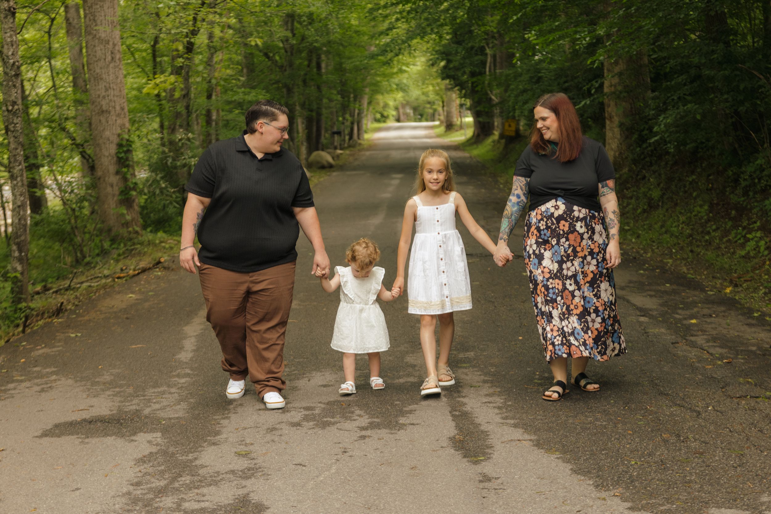 Family of four holding hands walking down a tree-lined path, two adults with two young girls in white dresses.