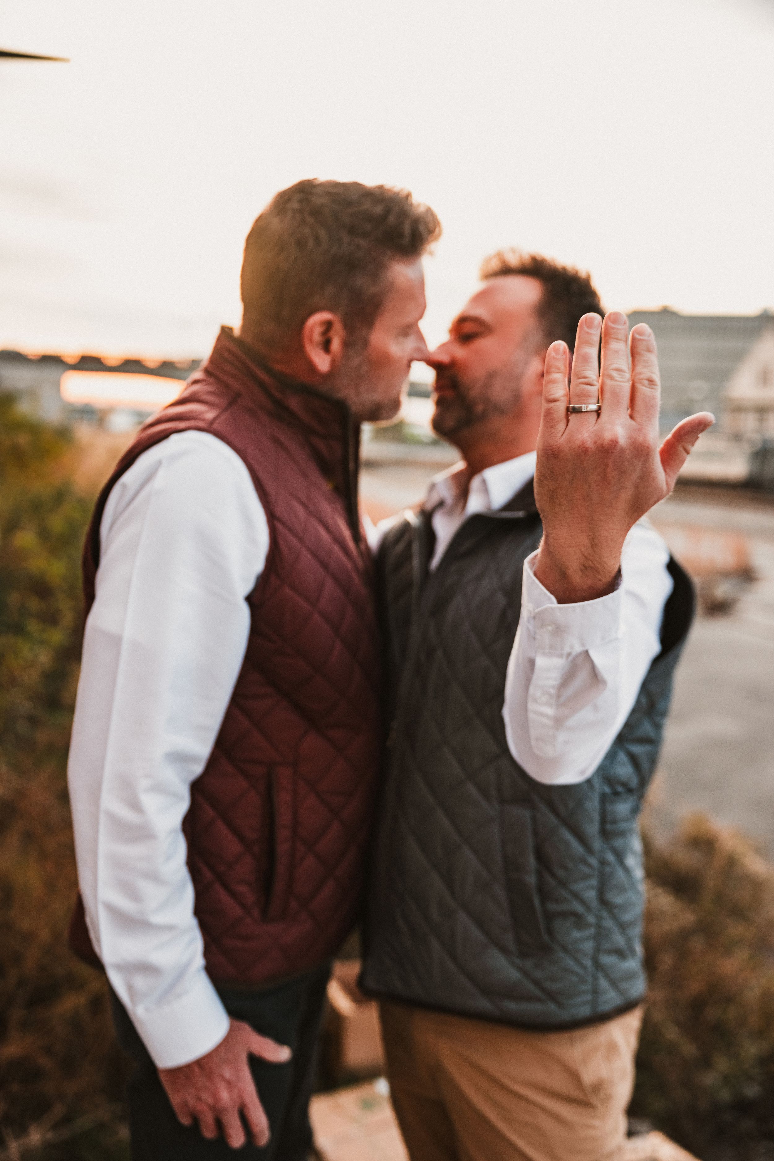 A couple in quilted vests sharing a romantic moment outdoors while showing off an engagement ring at sunset.