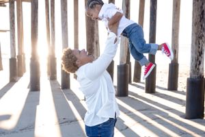 A parent lifts a small child wearing denim and pink shoes in the air while sunlight streams through wooden posts.