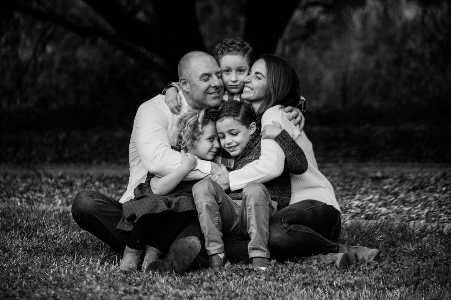 Intimate black and white family portrait showing loving embrace on grass with dark background.