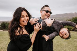 A family in dark clothing smiles and plays together while standing in a grassy field with hills in the background.
