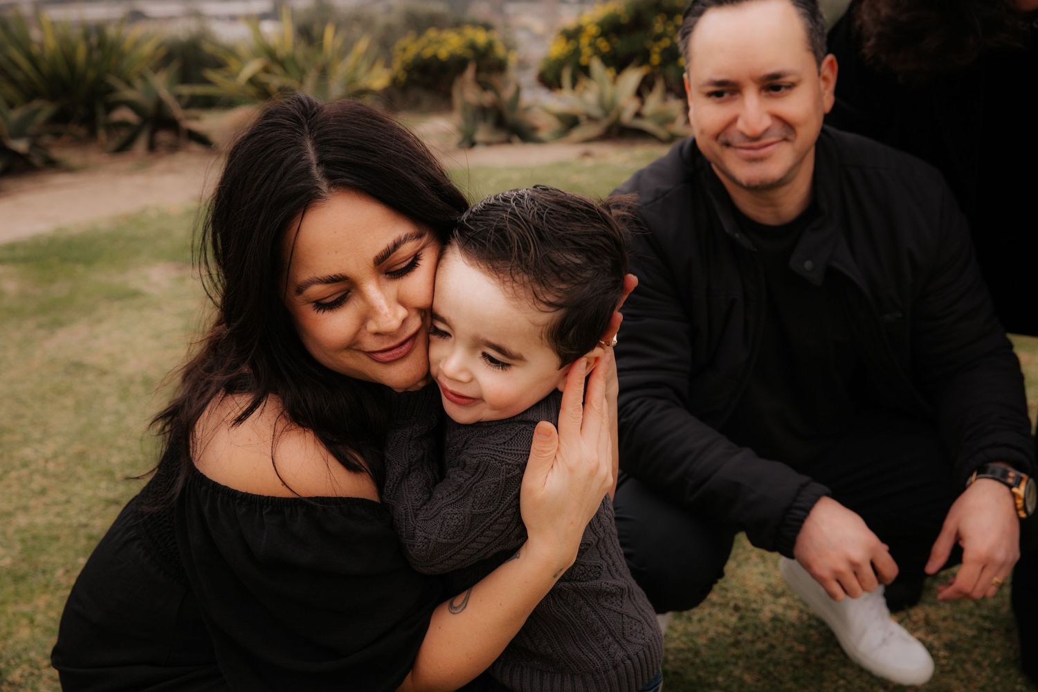 A family wearing black sits together outdoors on grass during a casual photo session in warm lighting.