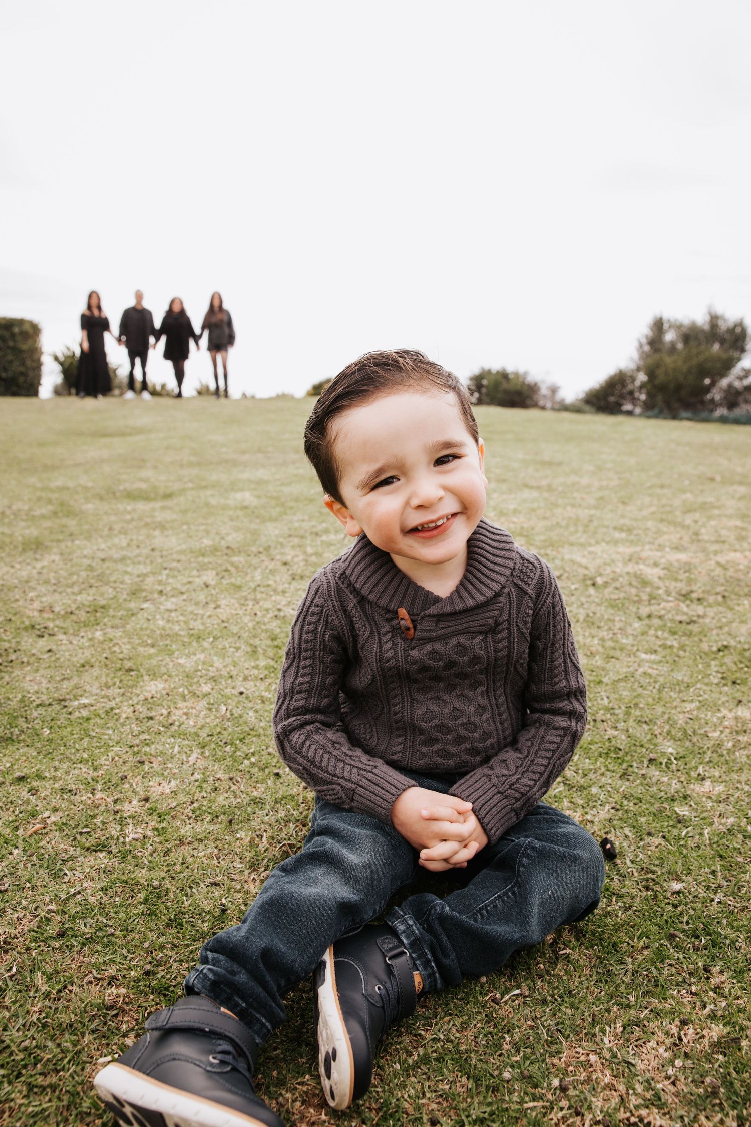 Child sits on grass in a brown knit sweater and jeans while a group stands in background on a grassy hill.