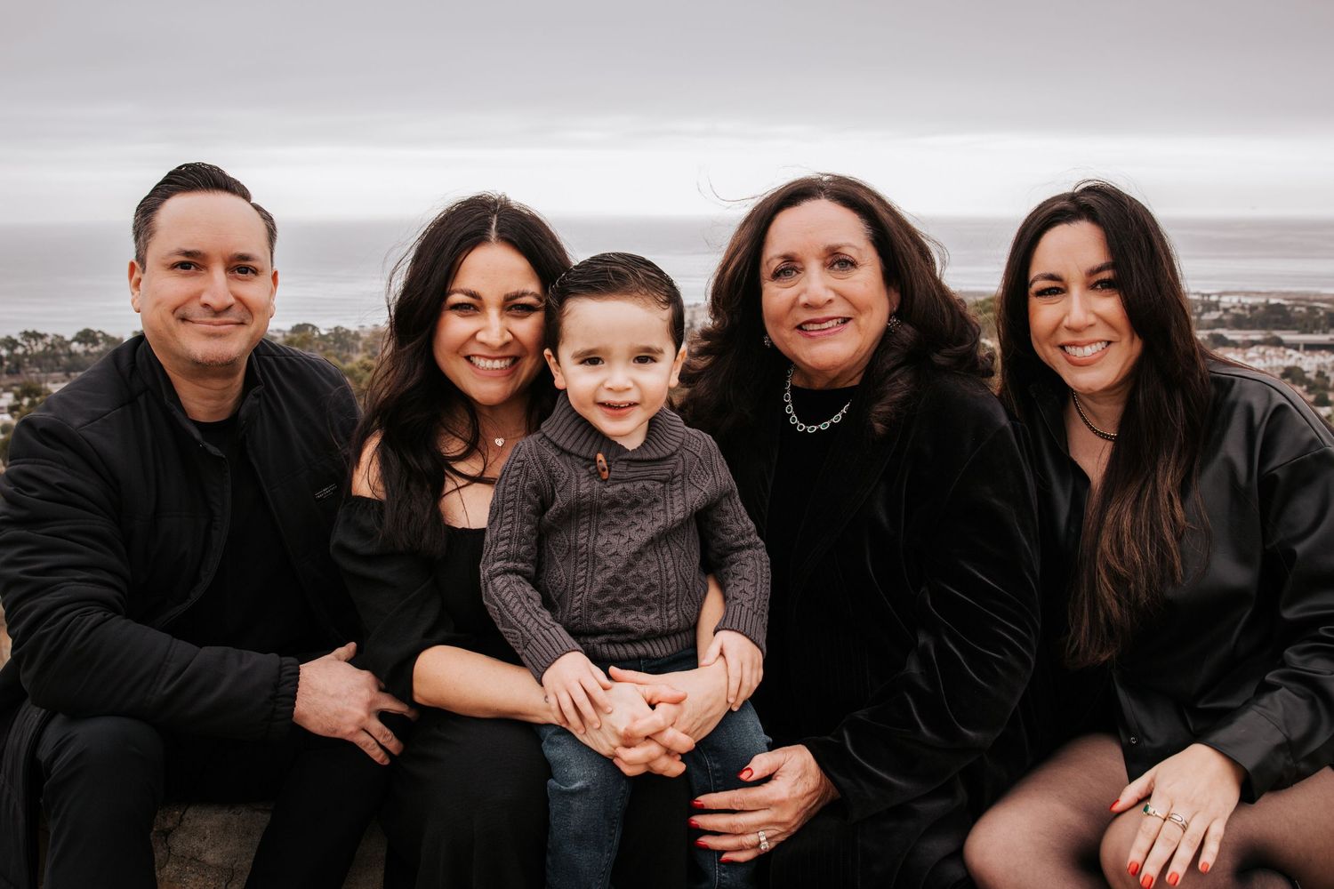 A family of five smiling together while sitting outdoors on a cloudy day at the coast.