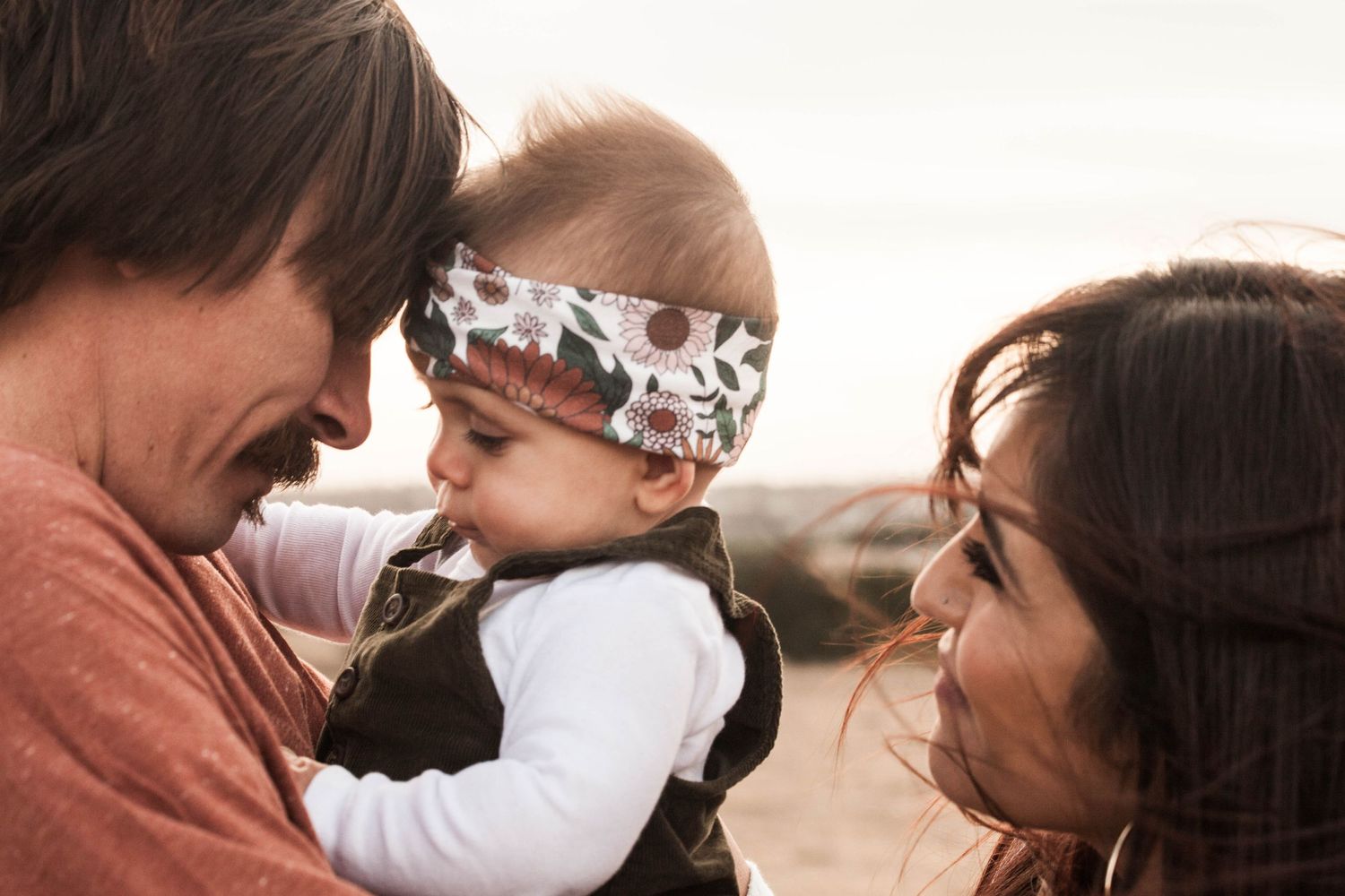 Three people share a tender outdoor moment in natural lighting with an infant wearing a patterned headband.