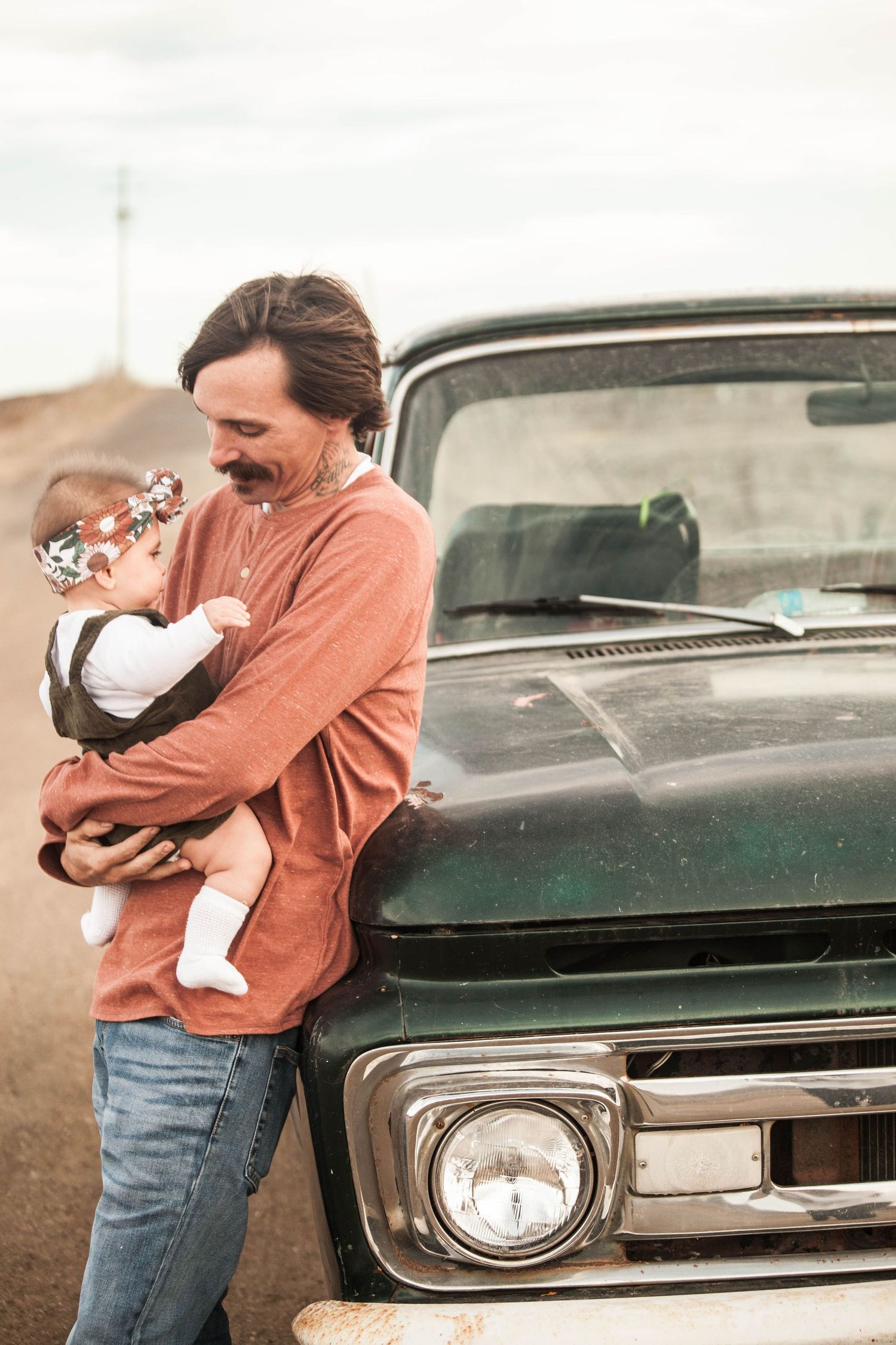 Person wearing orange sweater holding baby near vintage green truck in rustic outdoor setting.