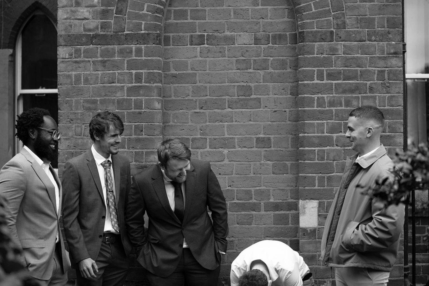 A group of people in formal attire stand against a brick wall in a black and white photograph.