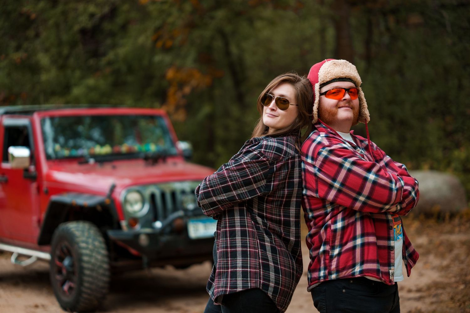 People in plaid shirts pose playfully near a red Jeep against a wooded autumn backdrop.