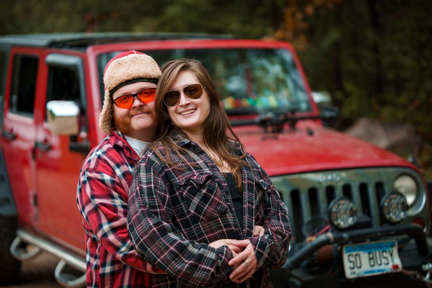 A couple sits together in autumn scenery near a red Jeep vehicle, enjoying a casual outdoor moment.