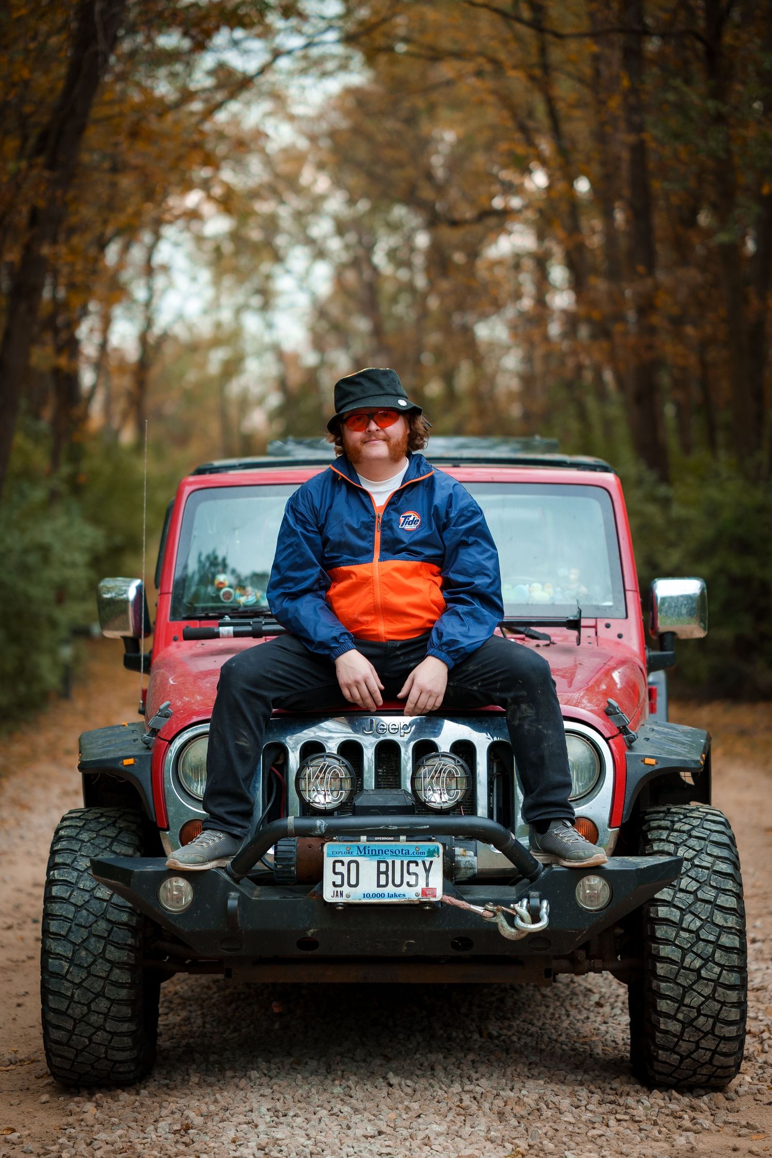 A red Jeep Wrangler with large off-road tires sits on a dirt path surrounded by autumn trees.