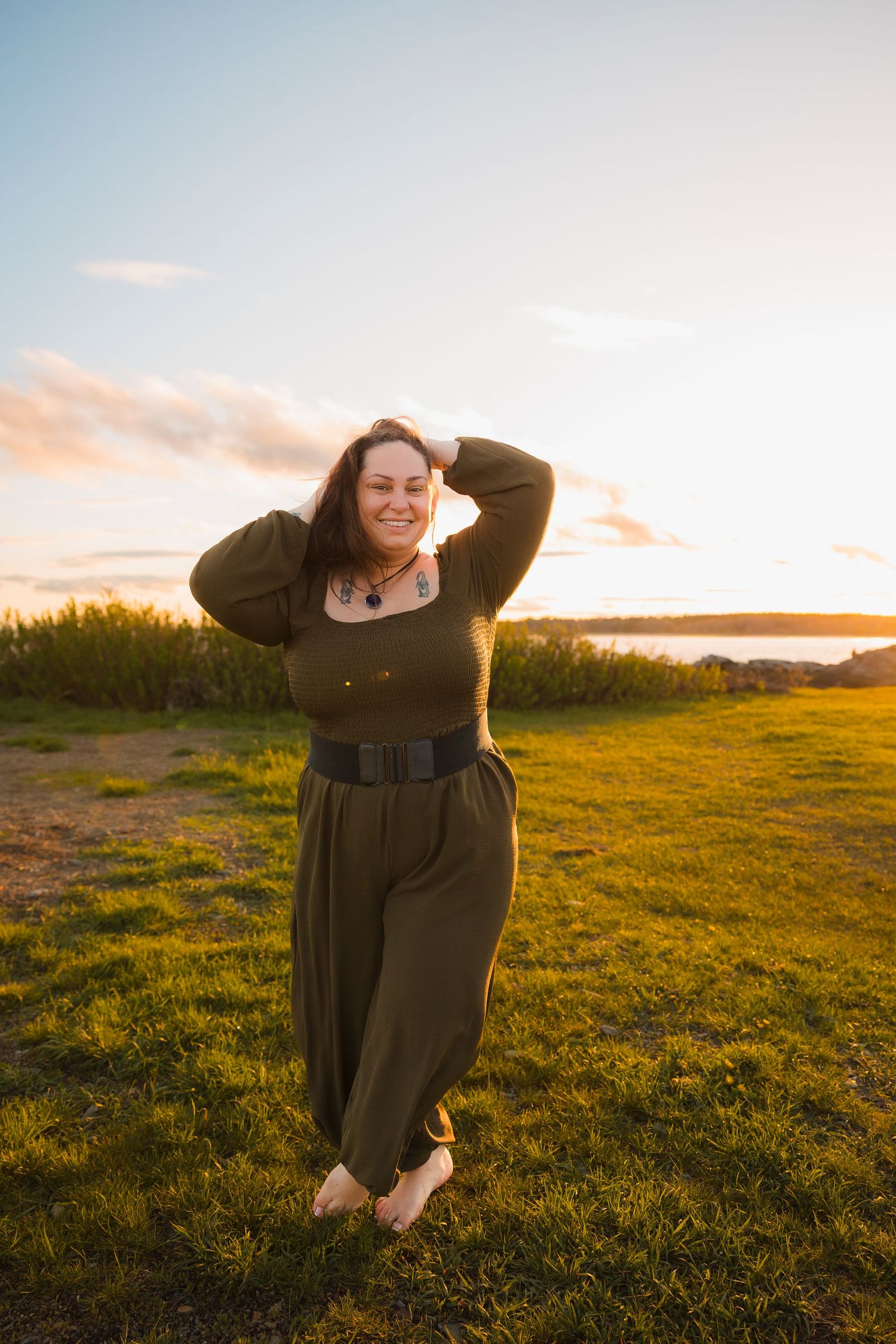 Person in an olive green dress poses against golden sunset light in grassy field.