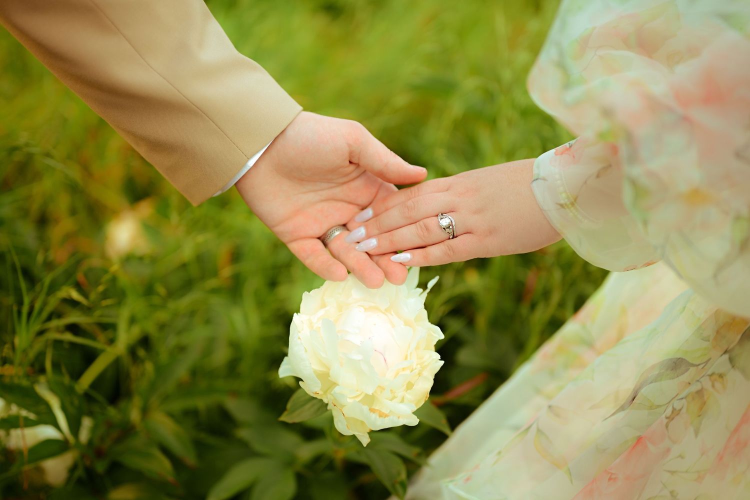 Two hands touching over a white peony flower, with a diamond engagement ring visible, set against a lush green garden backdrop.