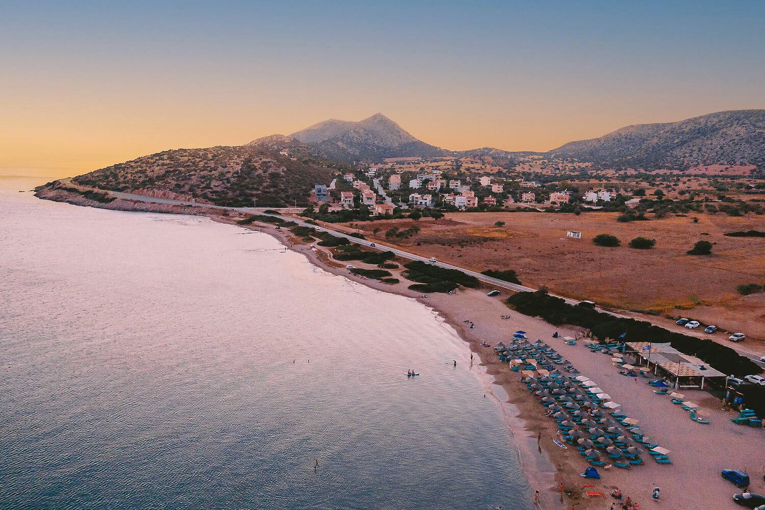 Aerial sunset view of a Greek coastal town with sandy beach, mountain backdrop, and calm blue waters.