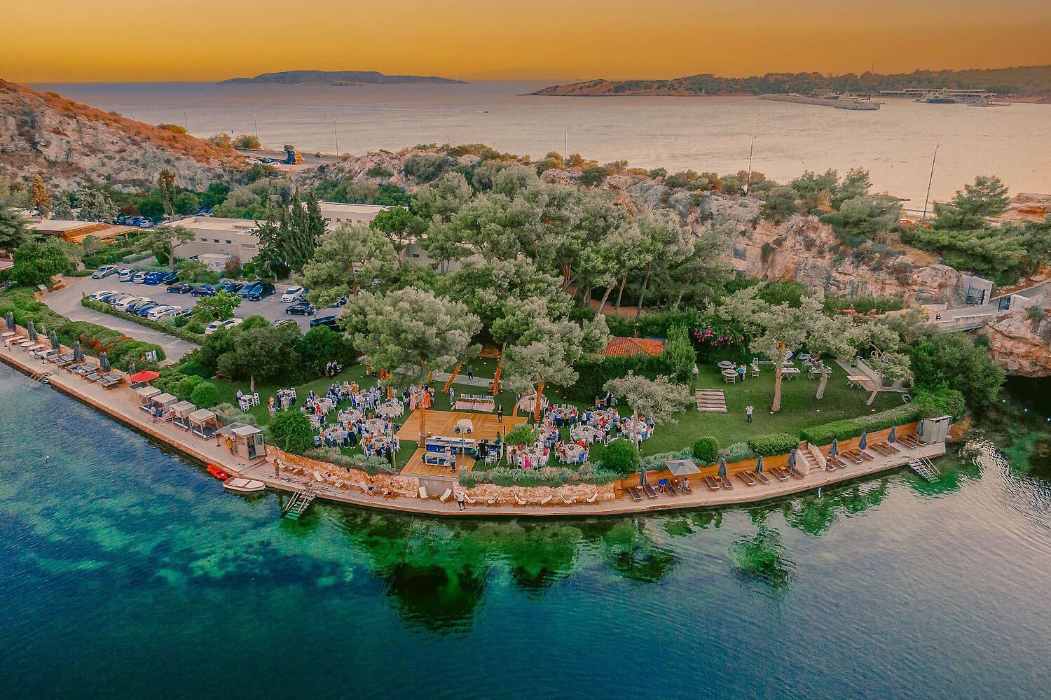 Aerial sunset view of a lush green peninsula resort surrounded by turquoise Mediterranean waters with mountains in the background.
