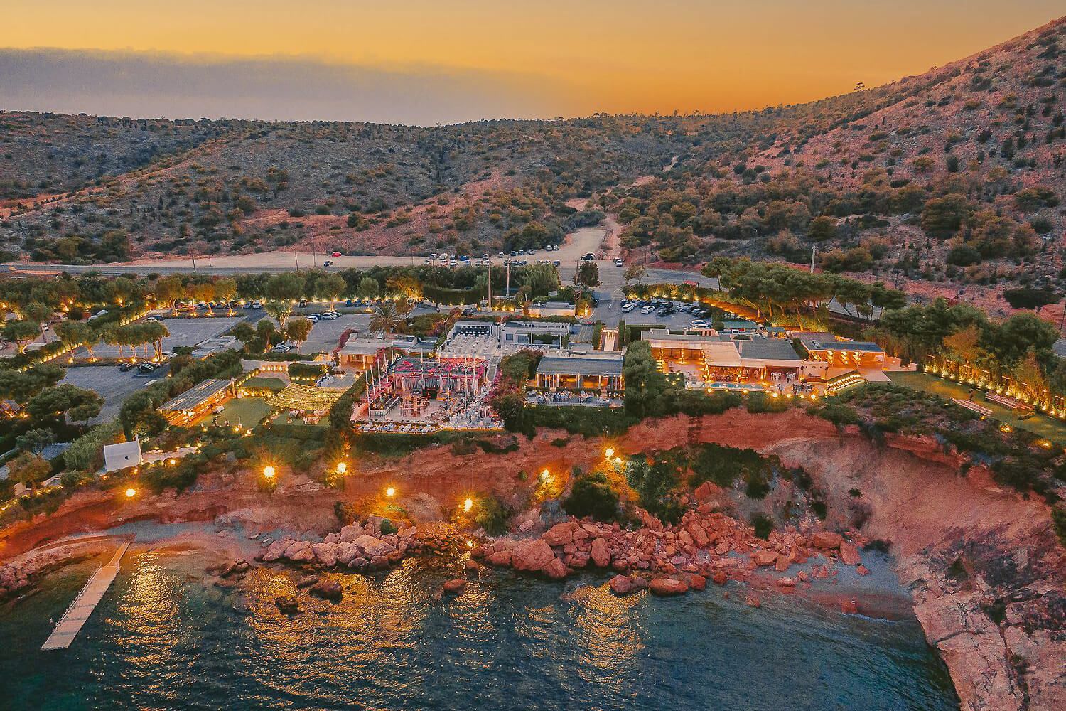Aerial sunset view of a luxury coastal resort with glowing lights nestled between rocky hills and calm blue waters.