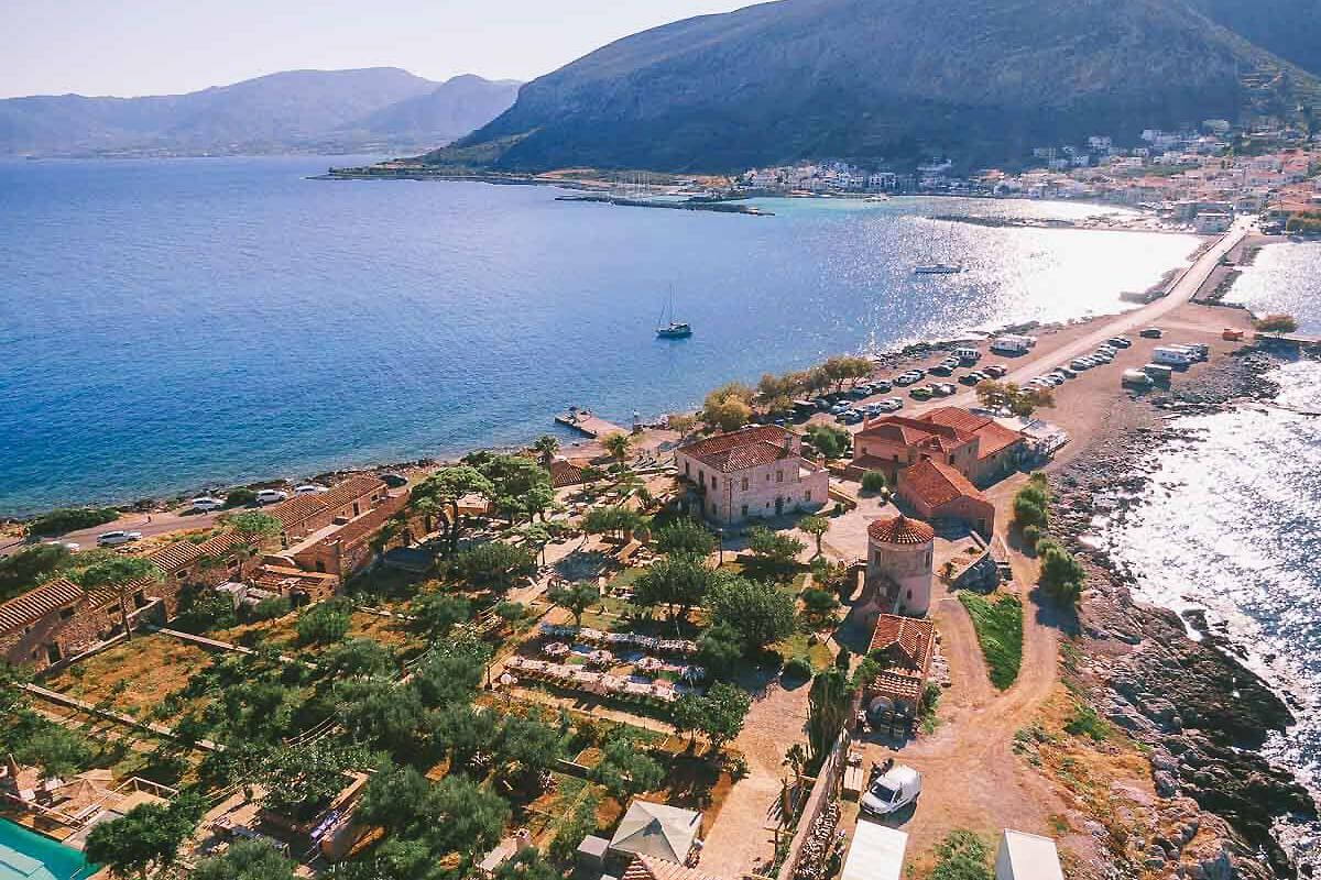 Aerial view of a Mediterranean coastal town with turquoise waters, historic buildings, and mountains in the background.