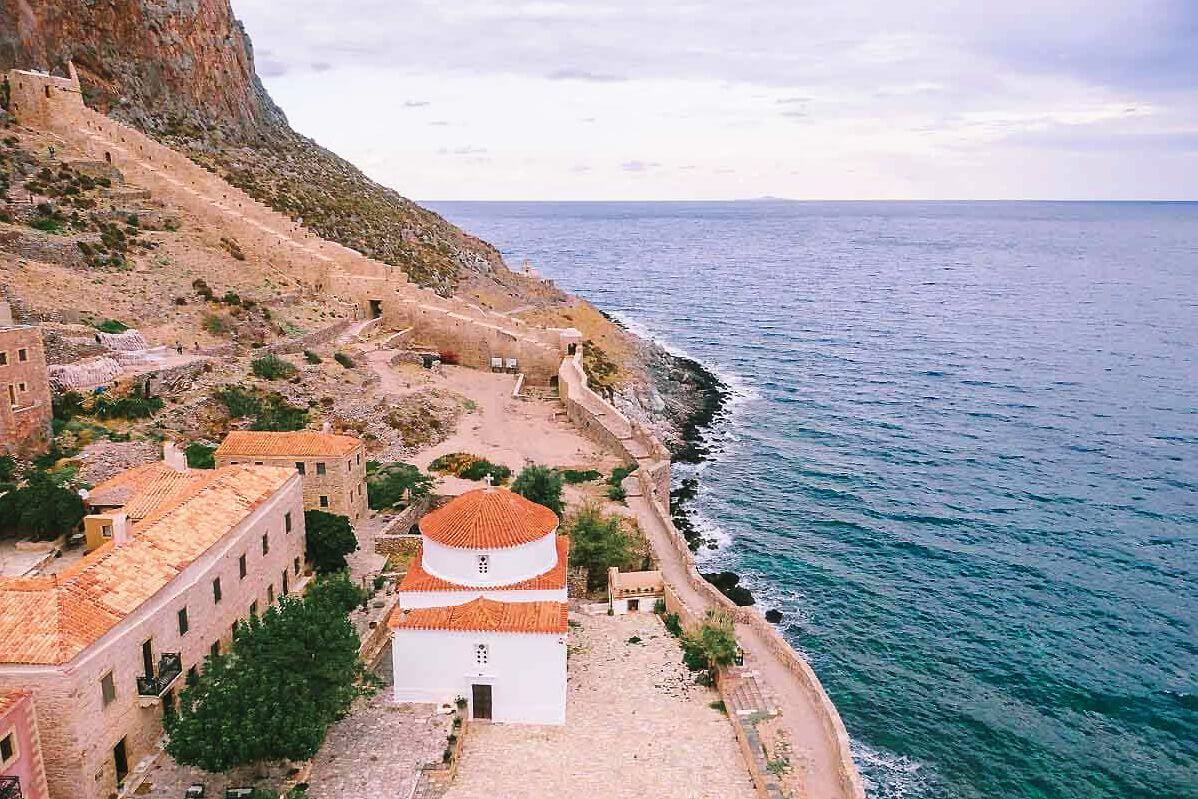Aerial view of a white-domed church beside turquoise Mediterranean sea, with stone buildings and cliffs in Greece.