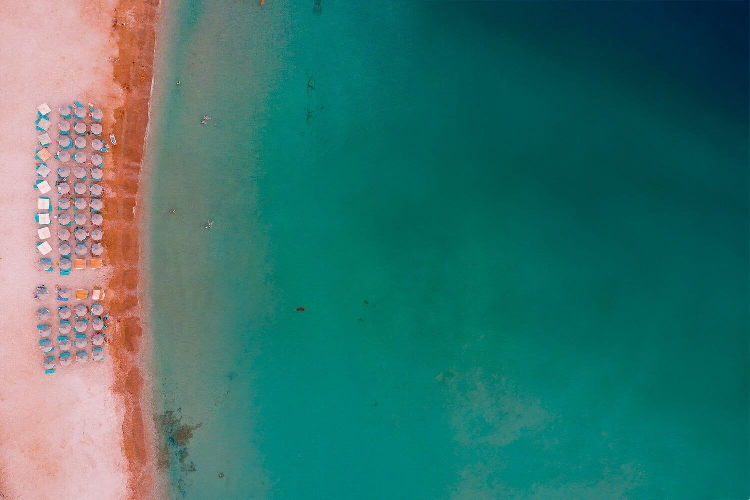 Aerial view of a turquoise beach with rows of umbrellas on red-orange sand along the shoreline.