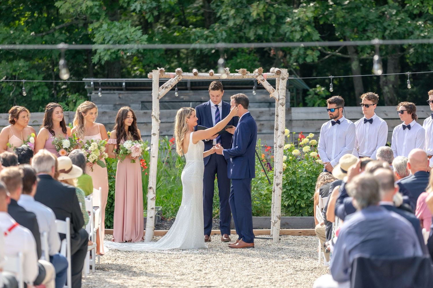 Bride and groom exchanging vows under a rustic wooden arch surrounded by wedding party outdoors.