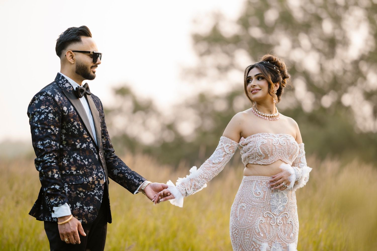 Couple in formal attire hold hands in a sunlit meadow wearing floral suit jacket and lace gown.