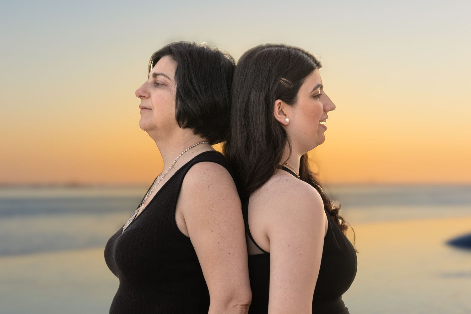 Two women in black dresses standing back-to-back against sunset ocean backdrop.