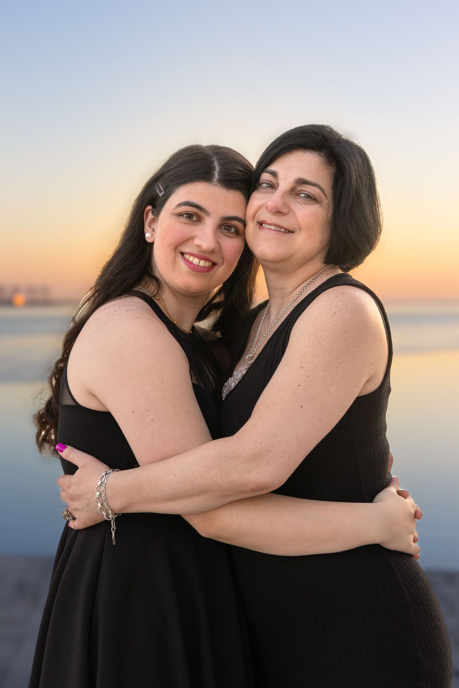 Two women in black dresses embracing and laughing by the ocean at sunset.
