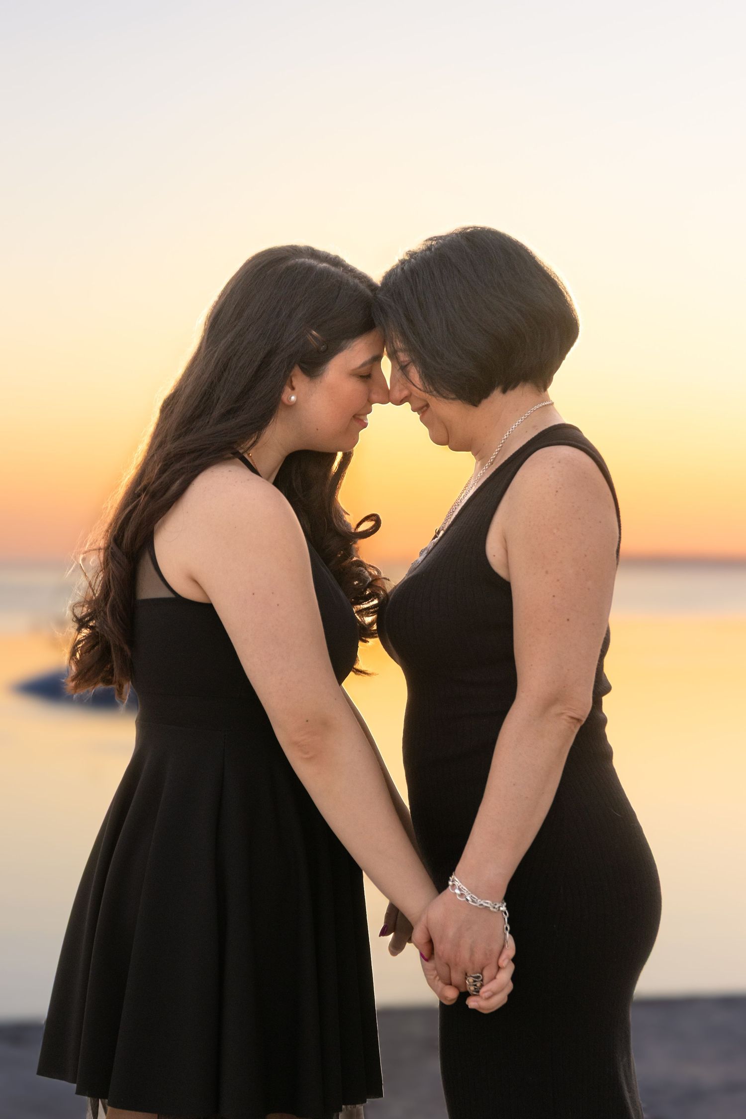 Intimate sunset portrait of two women in black dresses holding hands by the water.