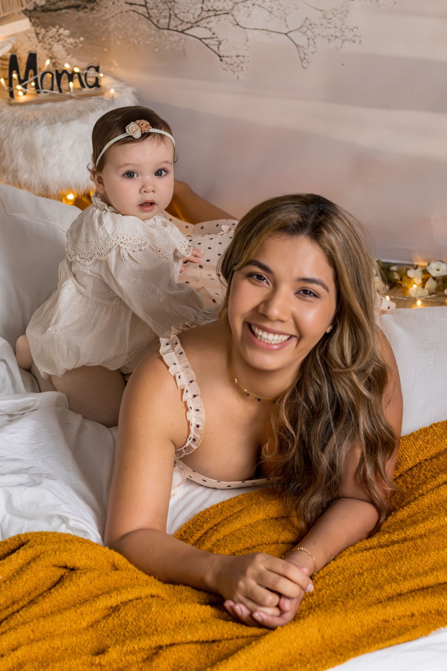 Smiling young mother lying on bed with adorable baby girl in white dress, cozy bedroom with warm fairy lights.